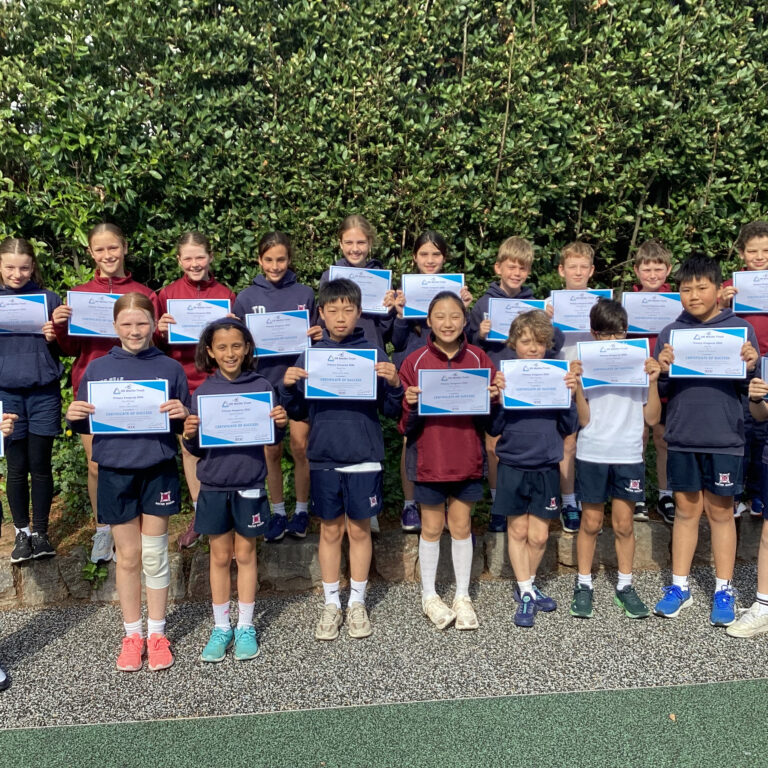 Group of children holding certificates outdoors in front of a green hedge, smiling at the camera.