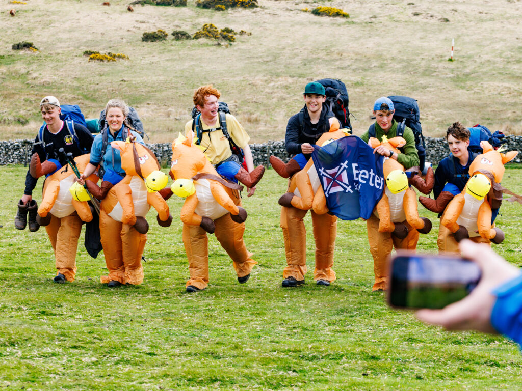 Group of people wearing orange inflatable dinosaur costumes with backpacks running together on a grassy field, smiling as they move forward.