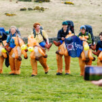 Group of people wearing orange inflatable dinosaur costumes with backpacks running together on a grassy field, smiling as they move forward.