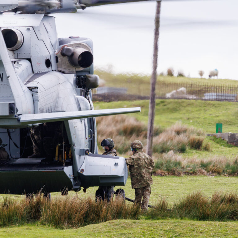Military transport helicopter on the ground with its rear cargo ramp open and soldiers nearby on a grassy field.
