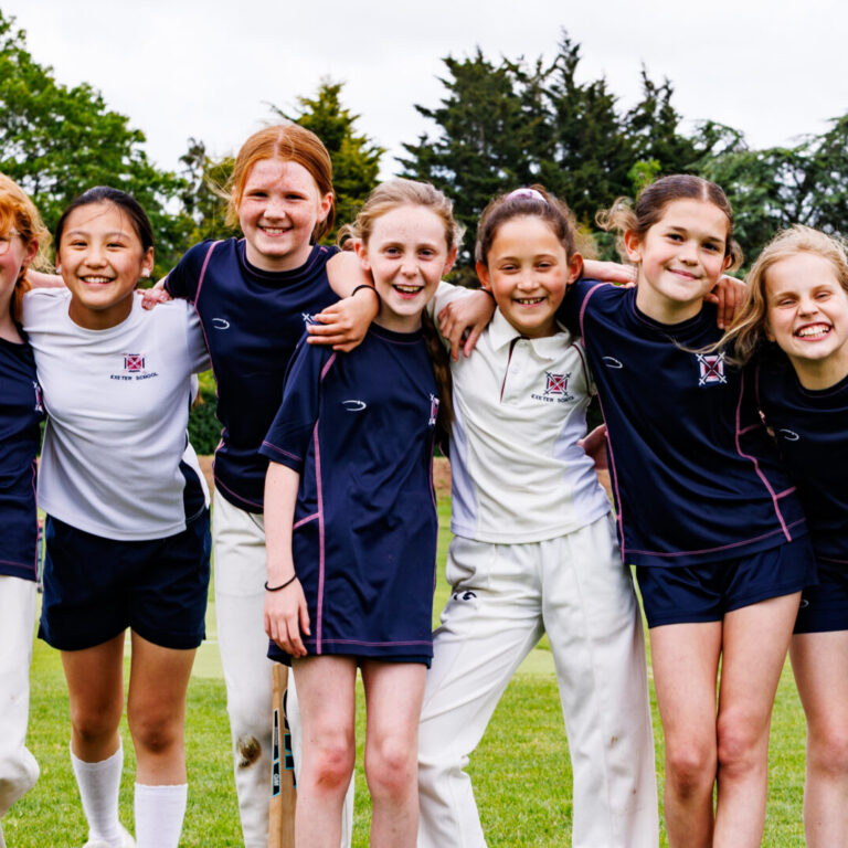 Youth cricket team posing together with bats and arms around each other on a grassy field, smiling at the camera.