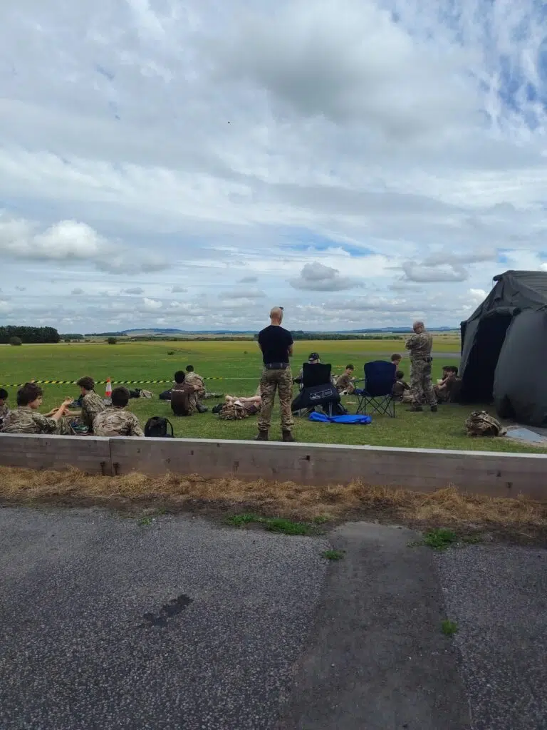 Group of soldiers in camo, some seated and standing on a grassy field near a gray tent, with a wooden barrier and cones in the foreground under a cloudy sky.