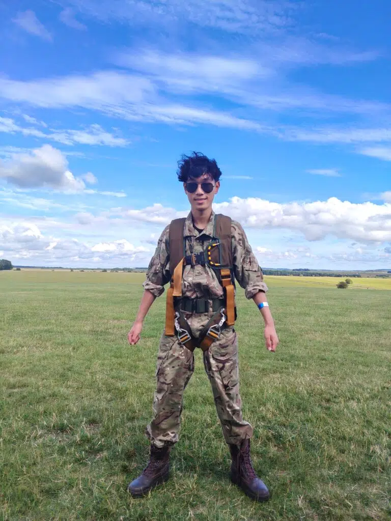 Person in camouflage military uniform wearing a parachute harness and sunglasses stands in a grassy field under a bright blue sky with scattered clouds, facing the camera.
