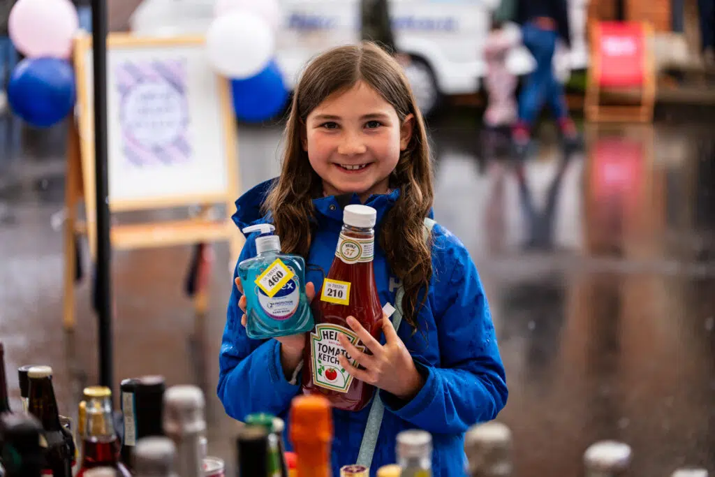 Smiling girl in a blue jacket holds a blue hand sanitizer bottle and a bottle of Heinz ketchup at a table covered with assorted bottles.
