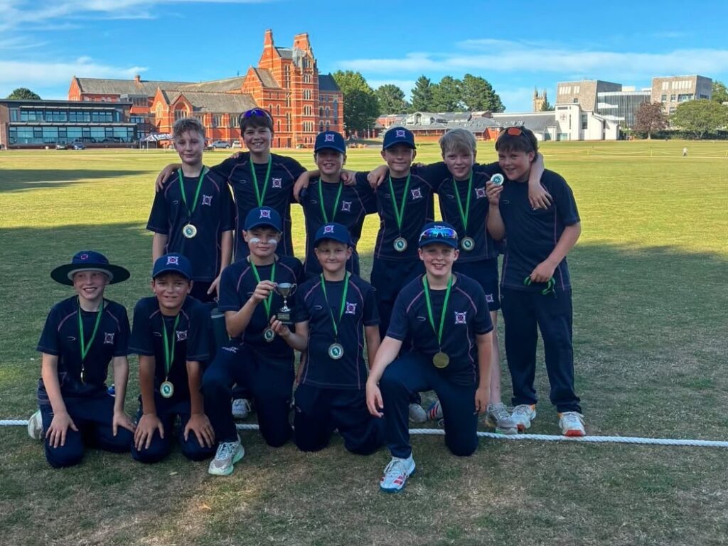 Eleven young athletes in matching black uniforms with green medals pose on a grassy field, one child holding a trophy, with a red-brick building in the background.