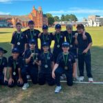 Eleven young athletes in matching black uniforms with green medals pose on a grassy field, one child holding a trophy, with a red-brick building in the background.