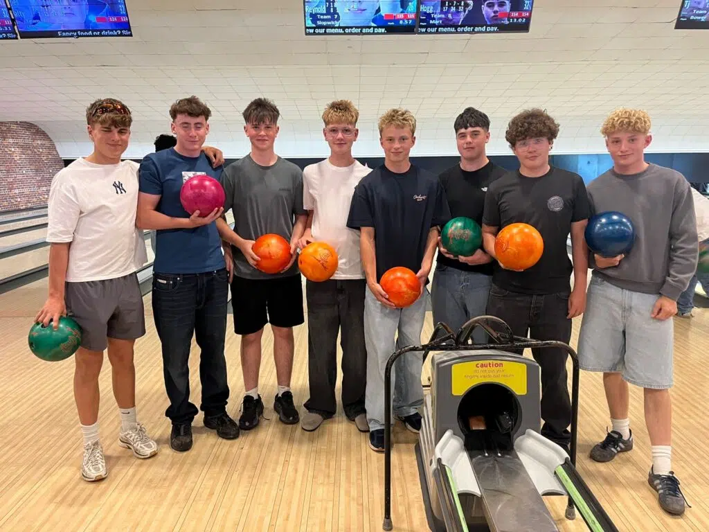 Nine young men posing with bowling balls in a bowling alley, standing side by side near the lane and ball return.