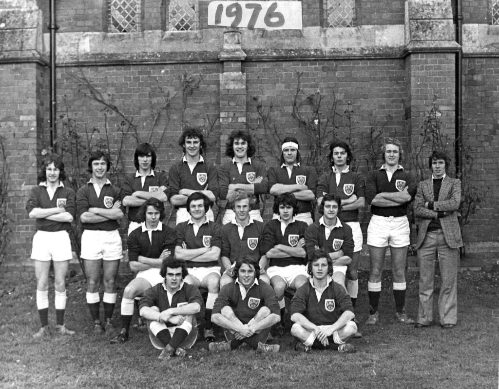 Group of football/rugby players in matching jerseys with crests, posing with arms crossed in front of a brick wall; 1976 banner above them (black-and-white photo).