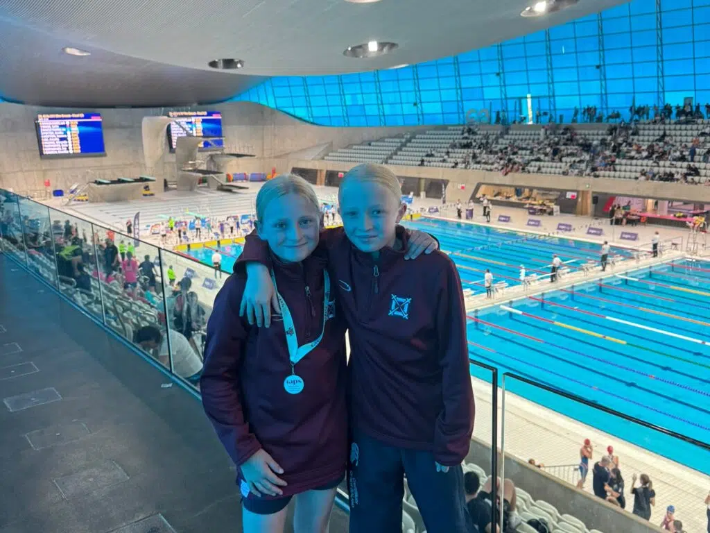 Two young swimmers in maroon jackets pose with medals at an indoor swimming pool competition venue. Behind them, spectators sit in the stands.