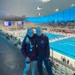 Two young swimmers in maroon jackets pose with medals at an indoor swimming pool competition venue. Behind them, spectators sit in the stands.