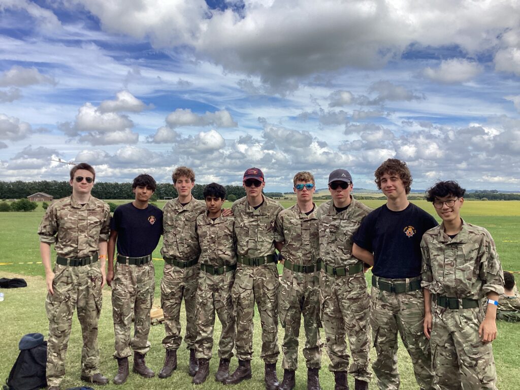 Group of young cadets in woodland camouflage uniforms posing for a photo on a grassy field under a cloudy sky.