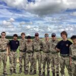 Group of young cadets in woodland camouflage uniforms posing for a photo on a grassy field under a cloudy sky.