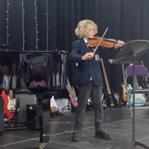 A young student in a blazer playing a violin on a stage, with a piano and guitars visible in the background.