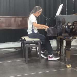 A young girl seated at a piano on a stage, playing the keys with sheet music on a stand nearby.