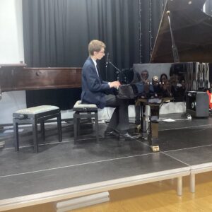 Young man in a blazer plays a glossy Yamaha grand piano on a stage with a microphone nearby.