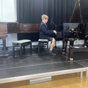 Young student in a navy blazer playing a black Yamaha grand piano on a stage with a microphone and curtains behind him.