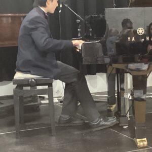 Profile of a young man in a dark suit playing a Yamaha grand piano on stage, microphone nearby.