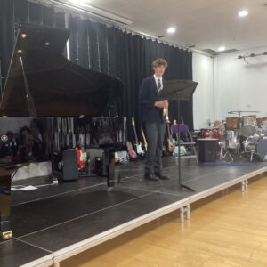 Young man in a suit stands on stage holding a trumpet beside a music stand with a grand piano to his left and various instruments in the background.