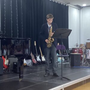 Young musician in a suit playing a saxophone on a stage with a music stand nearby and instruments in the background (piano, guitars, drums).