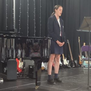 Young student singer on stage in a dark blazer, standing at a microphone with guitars in the background.