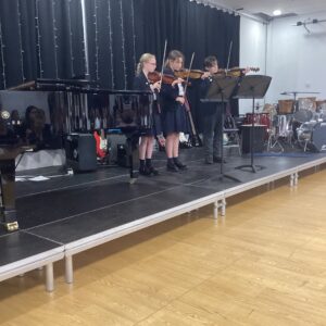 Three young violinists performing on a stage beside a grand piano, reading from sheet music stands while curtain backdrop hangs behind them