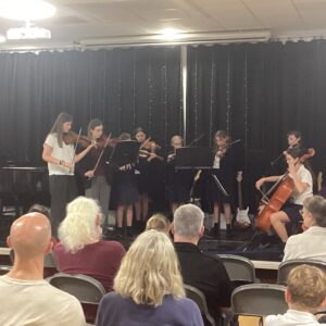 Young musicians playing string instruments on a stage with a black curtain; audience seated in front.