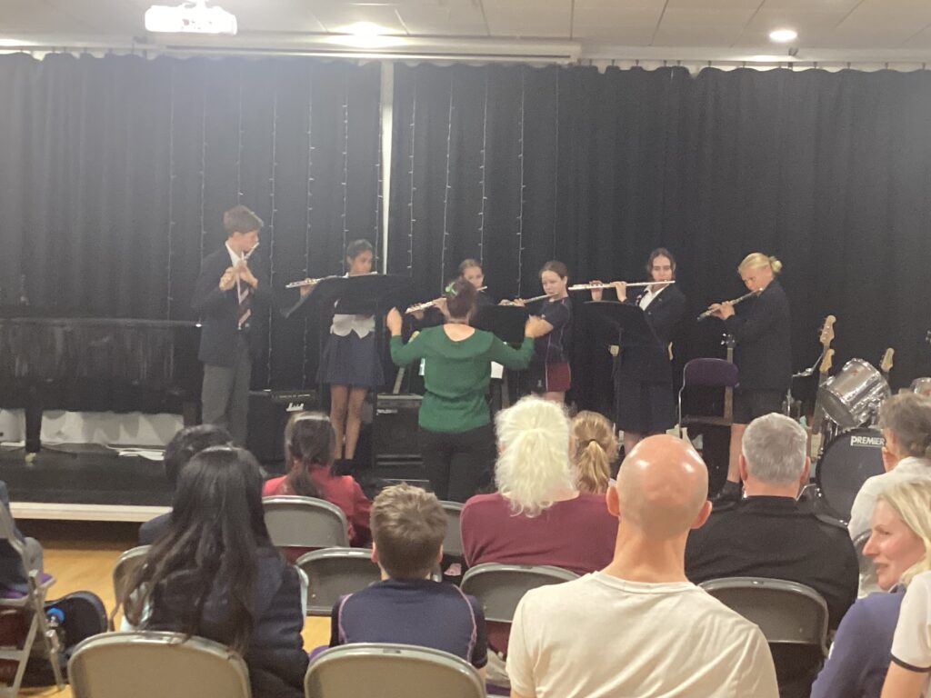 Conductor in green sweater leads a student band performing on a stage with flutes, behind a black curtain, while audience watches.
