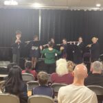 Conductor in green sweater leads a student band performing on a stage with flutes, behind a black curtain, while audience watches.
