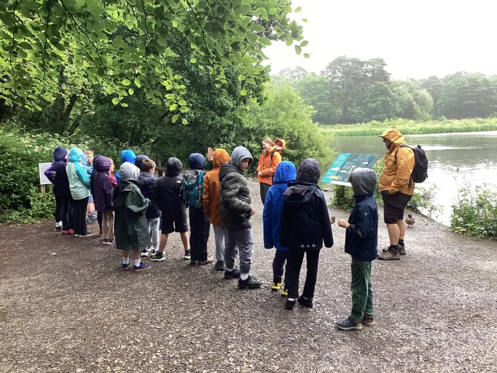 Group of children with backpacks on a gravel path by a lake, listening to a guide in orange jacket near an info board.