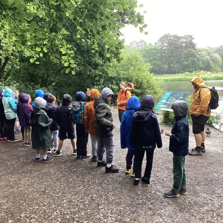 Group of children with backpacks on a gravel path by a lake, listening to a guide in orange jacket near an info board.