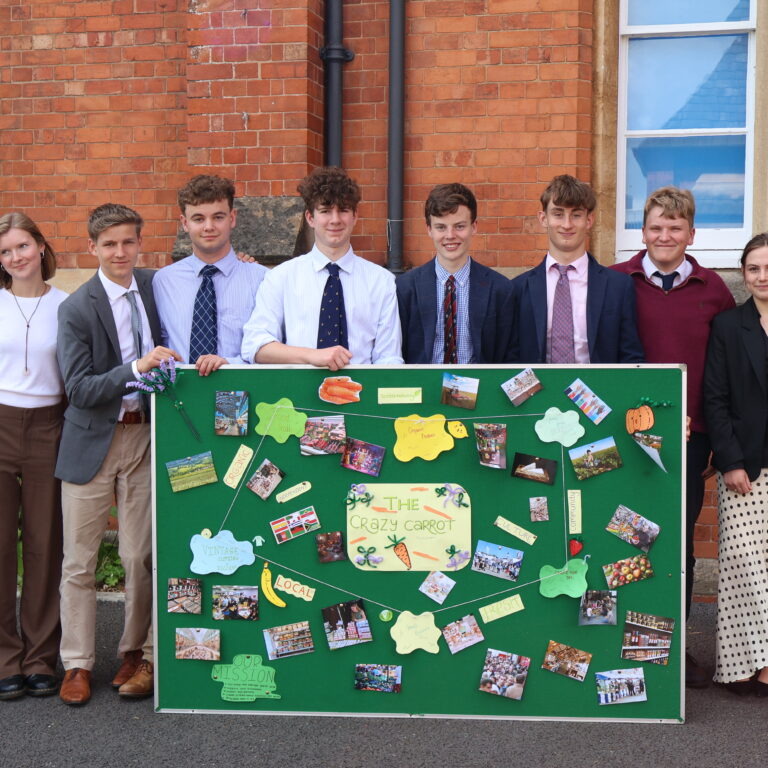 Ten students in smart attire pose behind a green presentation board covered with photos and notes outdoors by a red brick building.
