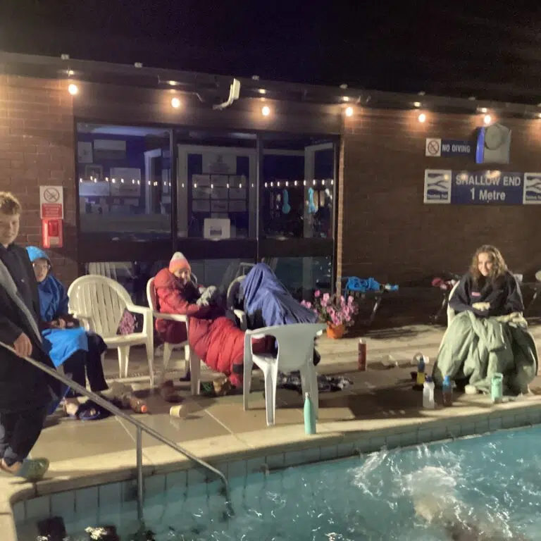 Group of people wearing jackets and hoodies sit around a pool at night under string lights, with a brick wall behind and pool in the foreground.