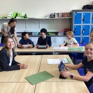 Group of smiling students sit at tables in a classroom as a teacher helps a student nearby.