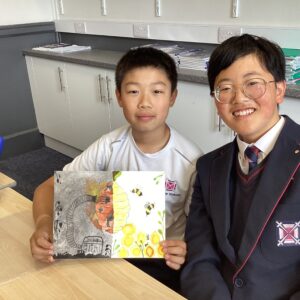 Two smiling students sit at a classroom table, presenting a colorful illustrated book with yellow hexagon shapes and nature drawings.