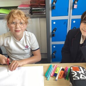 Two schoolchildren work at a desk drawing with colored markers; papers, a pencil case, and blue lockers are behind them in a classroom setting.