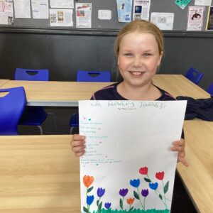 Smiling girl in a classroom holds up a large sheet of paper with a colorful tulip drawing in front of a bulletin board and blue chairs.