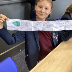 Girl in a blazer smiles while showing a long handwritten paper project to the camera in a classroom setting.