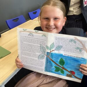 Smiling girl in a classroom holds open a colorful illustrated book with a tree drawing and text on the pages.