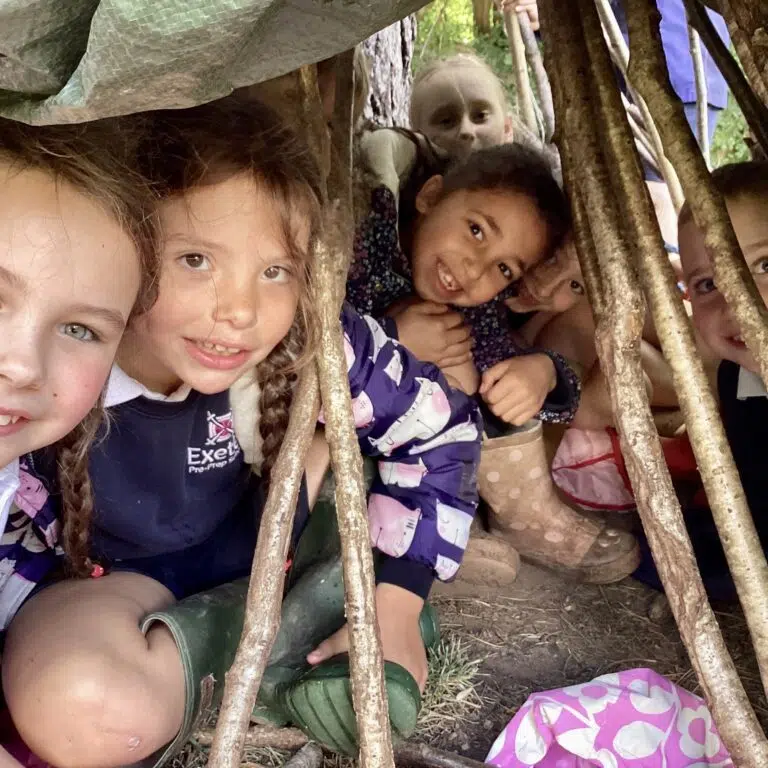 Group of smiling children peering through a handmade wooden fort structure outdoors.