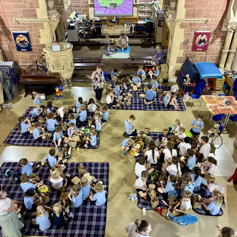 Group of children in blue uniforms sitting on plaid blankets in a large hall, doing crafts with bags and toys under adult supervision, with a stage area in back.