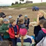 Group of children on a beach listening to a guide who is showing a tray of shells or rocks to the group.