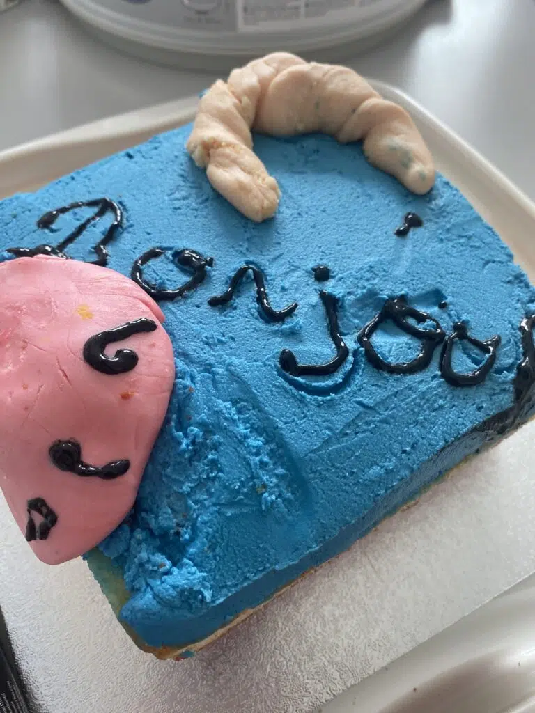 Blue frosted cake with a pink fondant finger sculpture resting on top and black icing writing (close-up).","Close-up of a blue frosted cake featuring a pink sugar finger and handwritten black icing.","Decorated cake: bright blue frosting, pink fondant finger, and black cursive text."