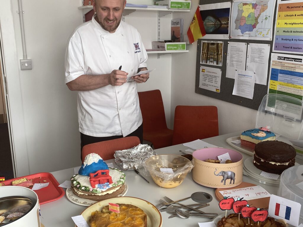 Chef in a white uniform stands beside a table full of cakes and pastries at a classroom celebration with Spanish flags on the wall.