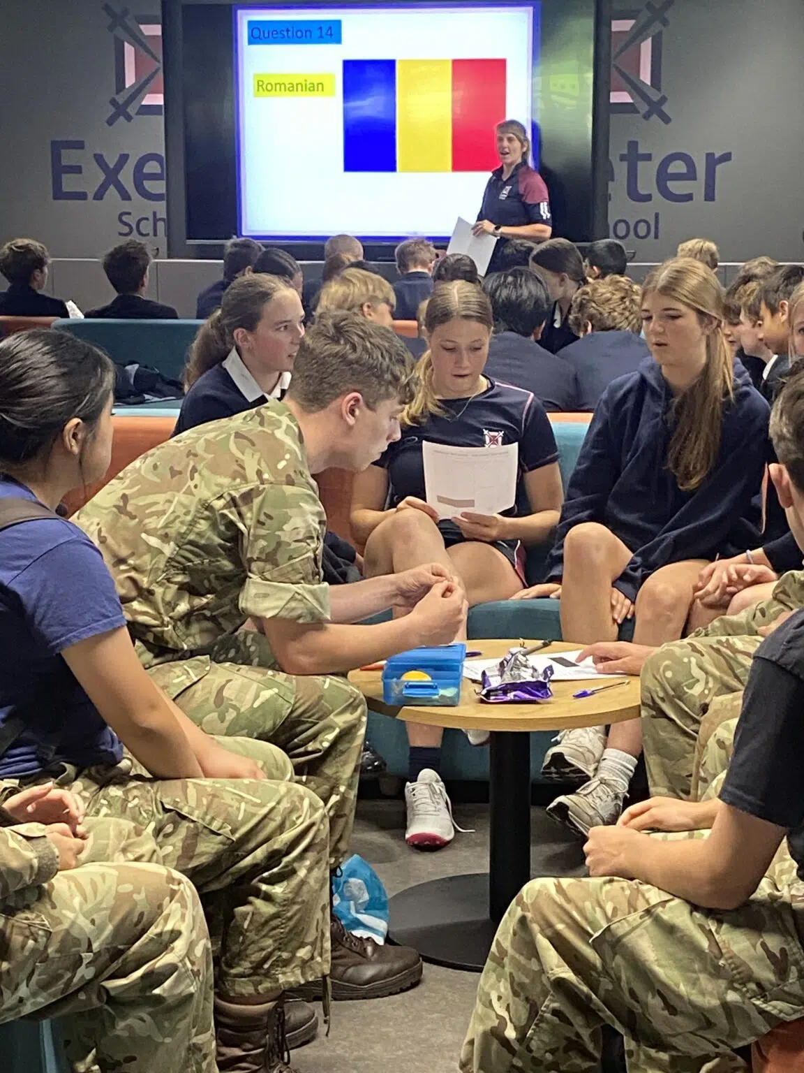 Group of students in camouflage uniforms sit around tables while a teacher presents a slide with Romanian flag colors on a screen at the front of the room.