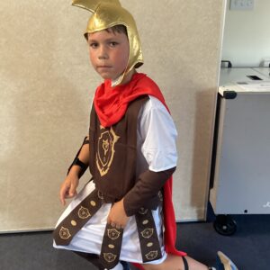 Boy in a knight costume kneeling on a dark floor, wearing a gold helmet, red cape, and brown tunic with crest.