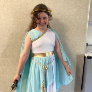 Girl in a light blue and white Greek-inspired costume with a gold belt and headband, posing with a staff or prop wand.