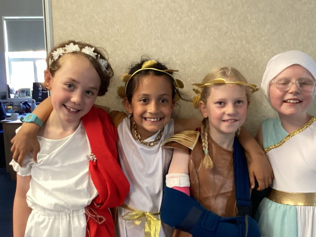 Four children in white and gold costumes posing together indoors, smiling with arms around each other in a classroom-like setting.