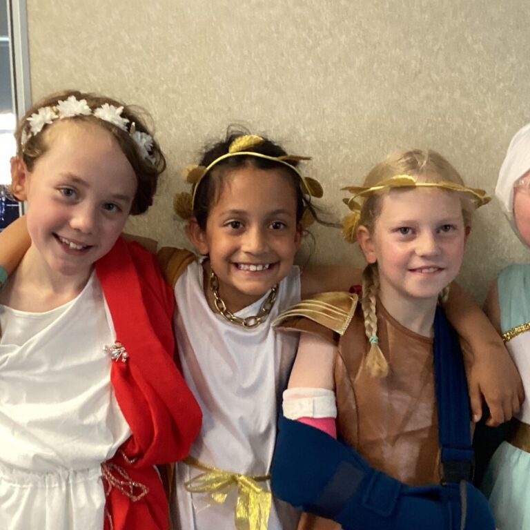 Four children in white and gold costumes posing together indoors, smiling with arms around each other in a classroom-like setting.