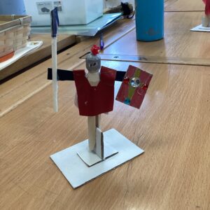 DIY humanoid figure made of popsicle sticks, red paper cape, and jeweled shield on a white base.