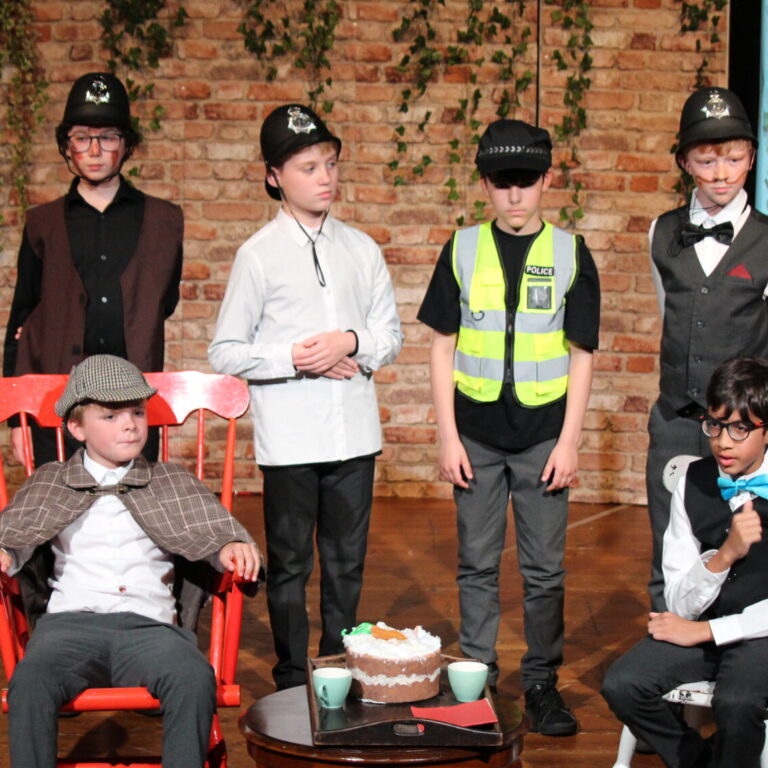 Children in a stage play wearing costumes (police hats, bow ties) gathered around a cake on a wooden table; brick wall and mural backdrop.